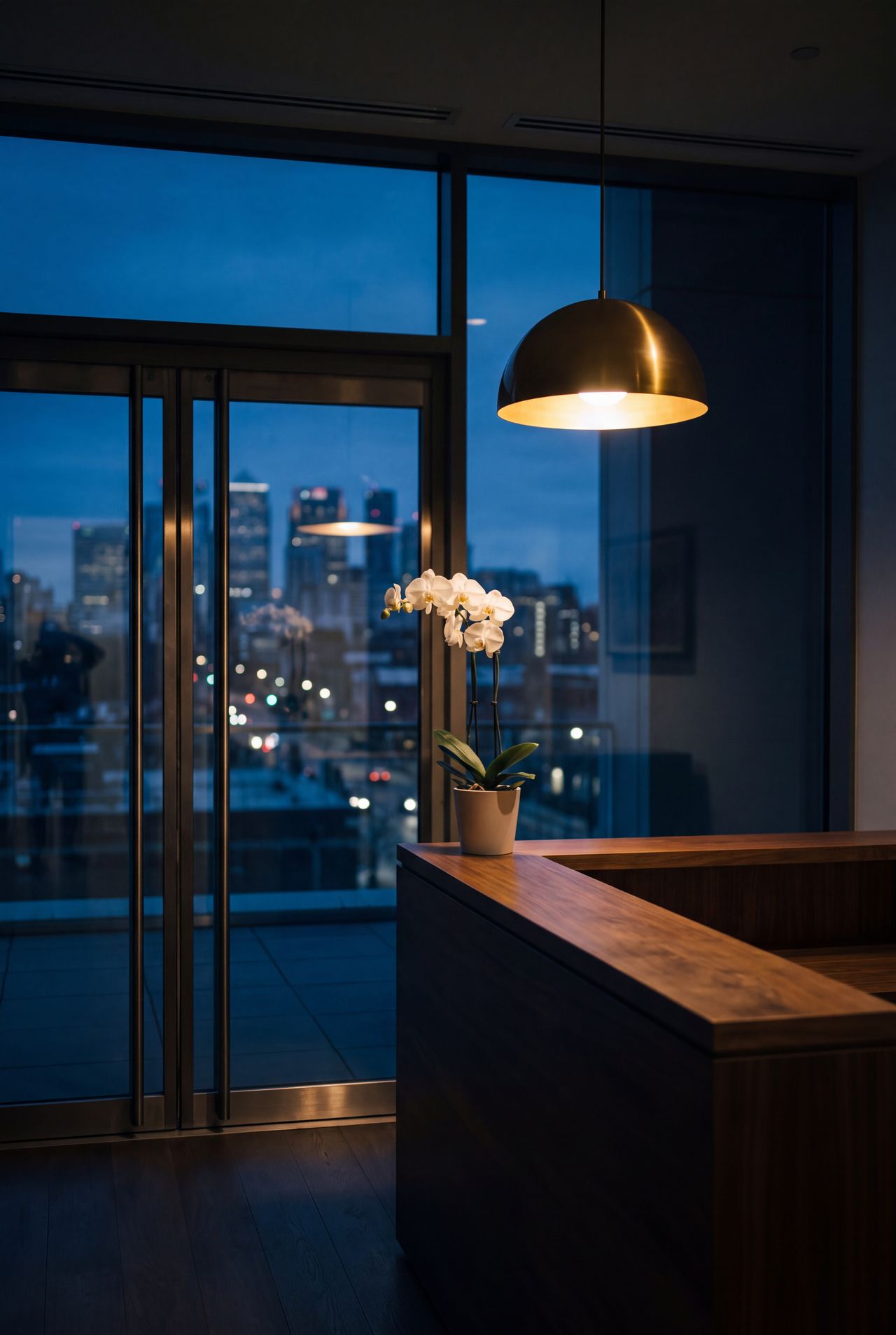 A reception at night, an orchid on the desk, a brass pendant lit, the city soft behind the glass