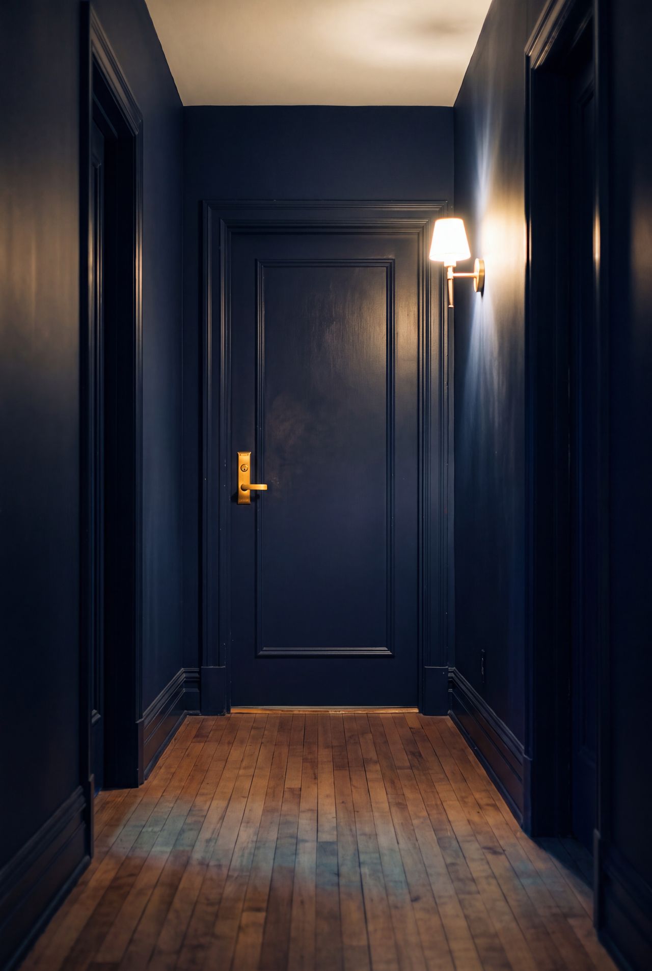 A navy hallway at dusk, a single brass door handle lit by a wall lamp