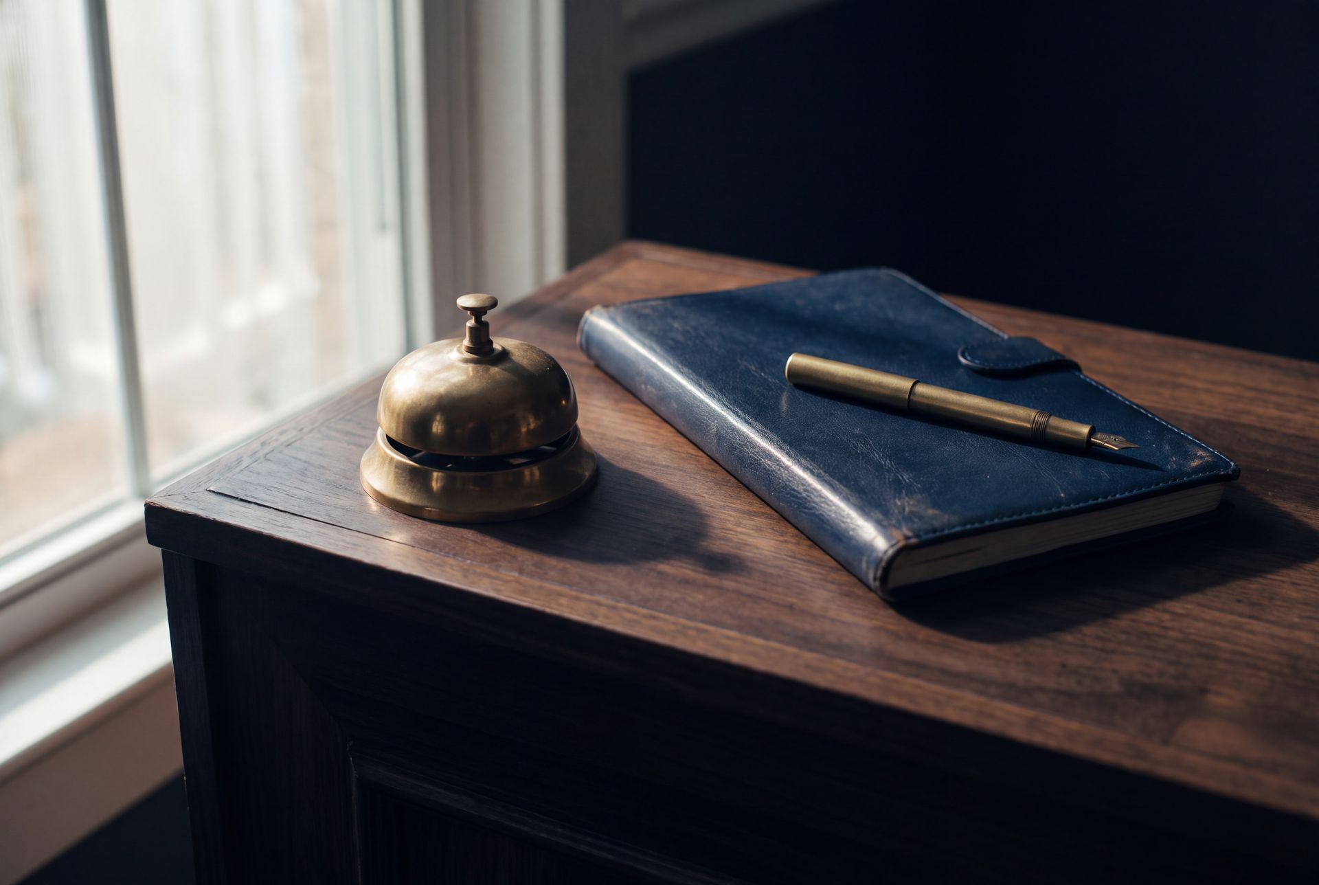 A brass service bell, a navy notebook and a pen on a walnut desk by a window