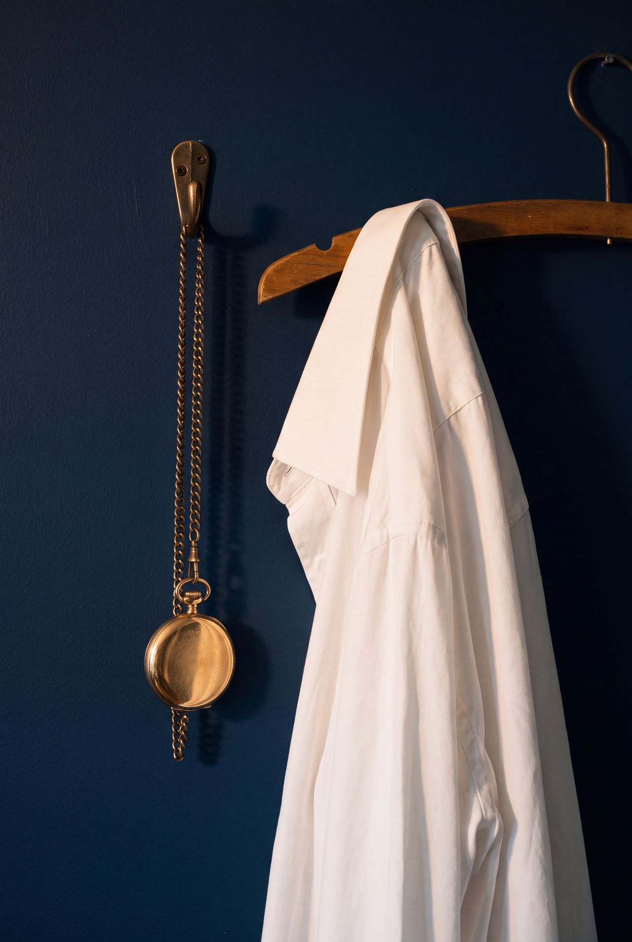 A pocket watch on a brass hook beside a white shirt on a navy wall