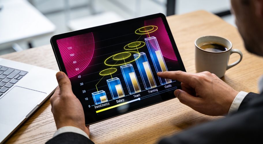 Person using a tablet to view a bar chart with categories for Investments, Salary, Team, Training, and Costs, considering whether to hire admin assistant or automate certain tasks, while enjoying a cup of coffee on the table.