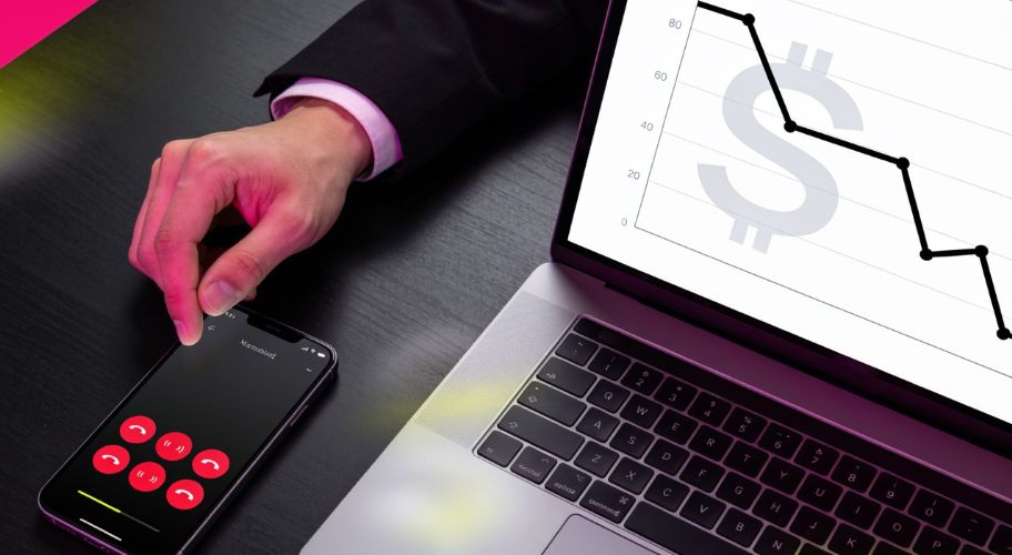 A person’s hand is near a smartphone with an active call, featuring a missed call text back notification, next to a laptop displaying a downward trending financial graph with a dollar symbol.