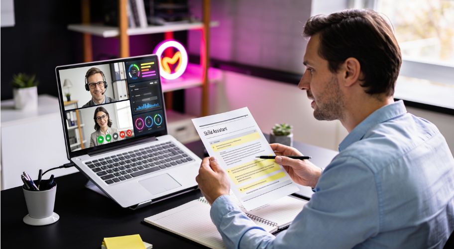 Man at a desk reviews a printed resume during a video call interview on his laptop with two people, with graphs and charts visible on the screen, possibly discussing virtual assistant cost as part of their hiring considerations.
