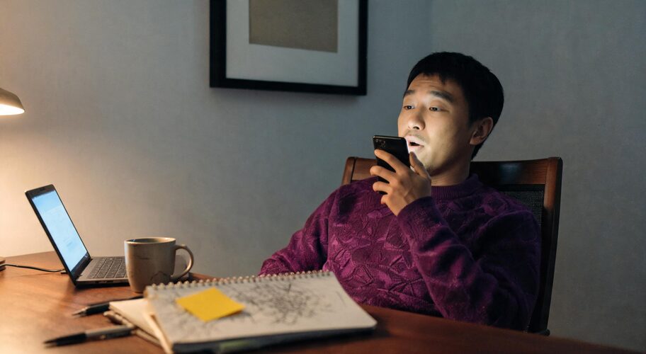 A person sits at a desk with a laptop, notebook, coffee mug, and sticky note, looking at a smartphone in a dimly lit room while using AI as a thinking partner.