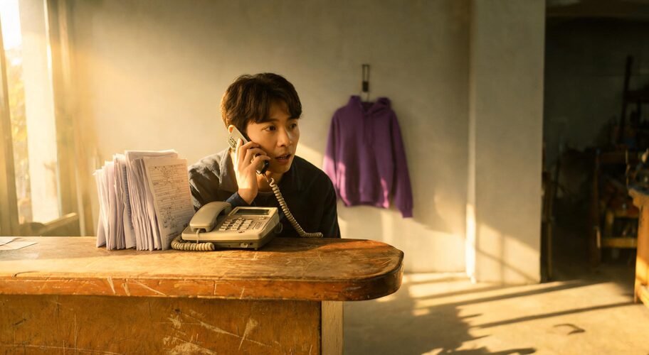 A small business owner stands behind a workshop counter in the late afternoon, holding a mobile phone to one ear while an office phone rings unanswered beside job dockets, highlighting the need to understand AI voice call disclosure rules.