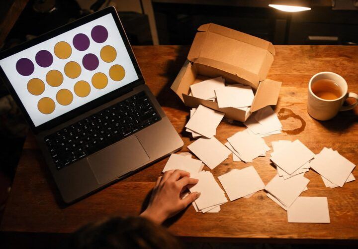 A person sorts blank cards on a wooden desk next to a laptop displaying colored dots, an open box with more cards, and a mug of tea—perhaps organizing information related to the Unsolicited Electronic Messages Act NZ.