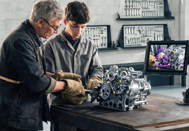 An older tradesman demonstrating AI training for apprentices in trades to a young apprentice working on an engine, with a tablet mounted nearby showing an augmented reality diagram of the engine components.