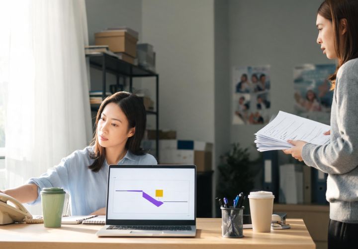 Two charity colleagues reviewing reports and workflow information for AI donor retention for NZ charities, with one holding a stack of papers beside a laptop displaying a chart.