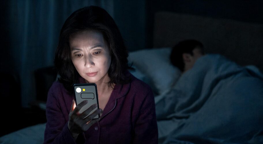 A host sits on the edge of a bed in a dark room, phone in hand and screen lighting their face, while a partner sleeps beside them, a late-night moment for AI for Airbnb hosts.
