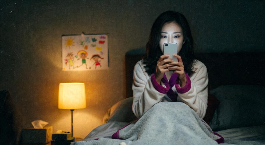 A woman using AI for family lawyers while sitting on the edge of a bed late at night, phone in both hands, lamp on, and a child's drawing stuck to the wall behind her.