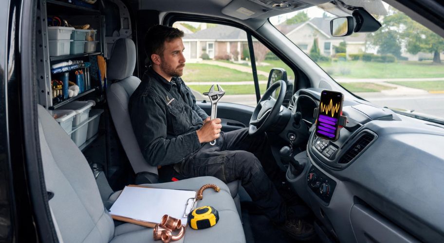 A man in work clothes sits in a van holding a wrench, with tools and equipment around him and a tablet on the dashboard displaying an AI receptionist for plumbers.