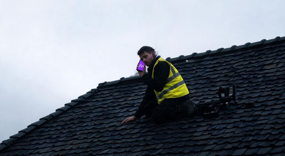 A person in a high-visibility vest kneels on a shingled roof while talking on a smartphone, serving as an AI consultant for tradies.